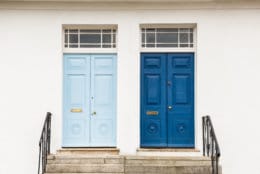 Two wooden blue, one dark blue, one light blue, Georgian style font doors to homes, against a white wall at the top of steps with metal banisters and windows at the top.
