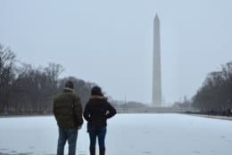 A couple takes in the first flakes late Saturday at the Reflecting Pool. The District and nearby suburbs could see upwards of 8 inches of snow by late Sunday. (WTOP/Dave Dildine)