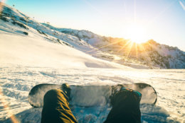 Snowboarder sitting at sunset on relax moment in french alps ski resort - Winter sport concept with adventure guy on top of mountain ready to ride down - Legs view point with teal and orange filter