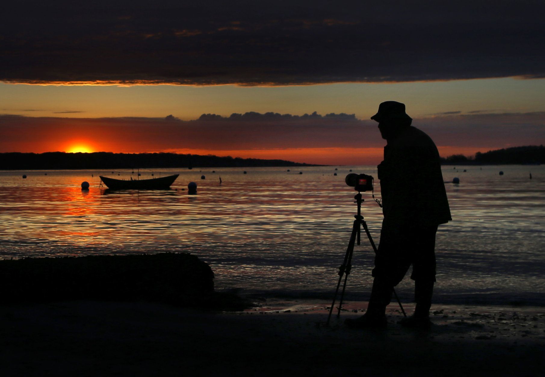 A brief appearance by the sun provides Chuck Johns with a dramatic backdrop as he photographs a dory moored off Willard Beach, Friday morning, May 17, 2019, in South Portland, Maine. The sun has been an infrequent visitor to the state in recent weeks. Another round of rain showers are expected to move in later in the day. (AP Photo/Robert F. Bukaty)