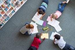 Elementary children lying on the floor and drawing at library. Top view of five multiethnic boys and girls in daycare house drawing on copybook. High angle view of group of kids drawing with colorful pencils on floor.