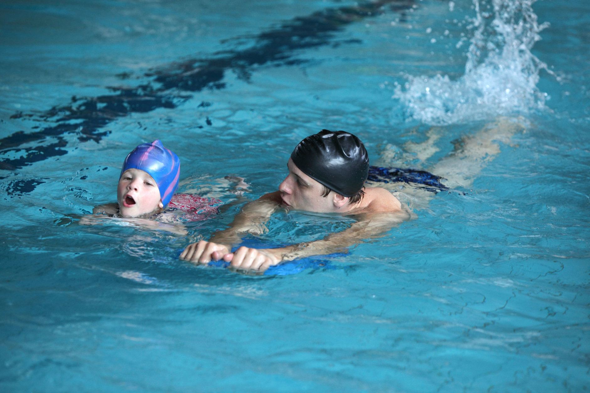 Close up of swimming lesson - child practicing flutter kick with kick board with instructor in indoor swimming pool - front view
