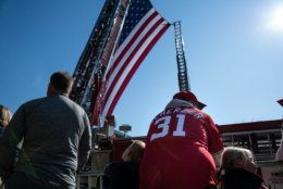 A Washington Nationals fan awaits the start of the parade on Constitution Avenue, beneath a behemoth American flag hoisted by two D.C. fire engines. (WTOP/Alejandro Alvarez)