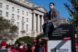 A Nationals fan sits atop a sign for the Smithsonian Museum of American History on Constitution Avenue. Fans mounted signs, ledges, power boxes and, on occassion, climbed street lights for a view above the dense crowd. (WTOP/Alejandro Alvarez)