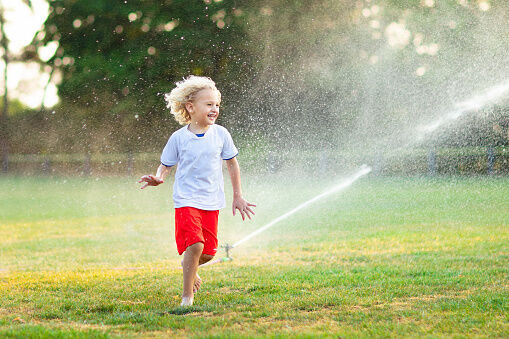 Kids play with water on hot summer day. Children with garden sprinkler. Outdoor fun. Boy and girl run on football field after training under water drops.