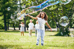 Adorable African American girl making soap bubbles while her little friends standing behind her and keeping eye on her, sunny public park on background