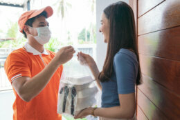 Delivery man in orange uniform delivering. Asian food in take away boxes to a woman customer at home. Deliver man wearing face mask handling food boxs give to female costumer