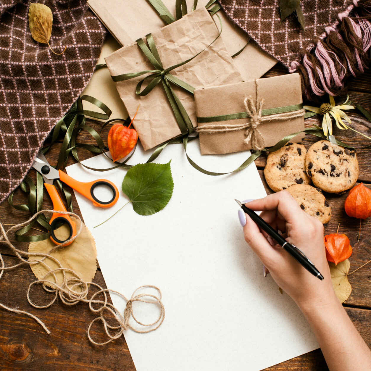 Autumn holidays and preparing presents, top view. Unrecognizable woman writing check list on wooden table near small wrapped gifts, chocolate cookies, scissor and bands laying on warm cozy plaid