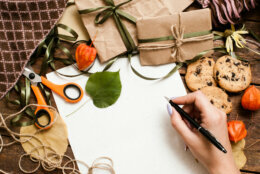 Autumn holidays and preparing presents, top view. Unrecognizable woman writing check list on wooden table near small wrapped gifts, chocolate cookies, scissor and bands laying on warm cozy plaid