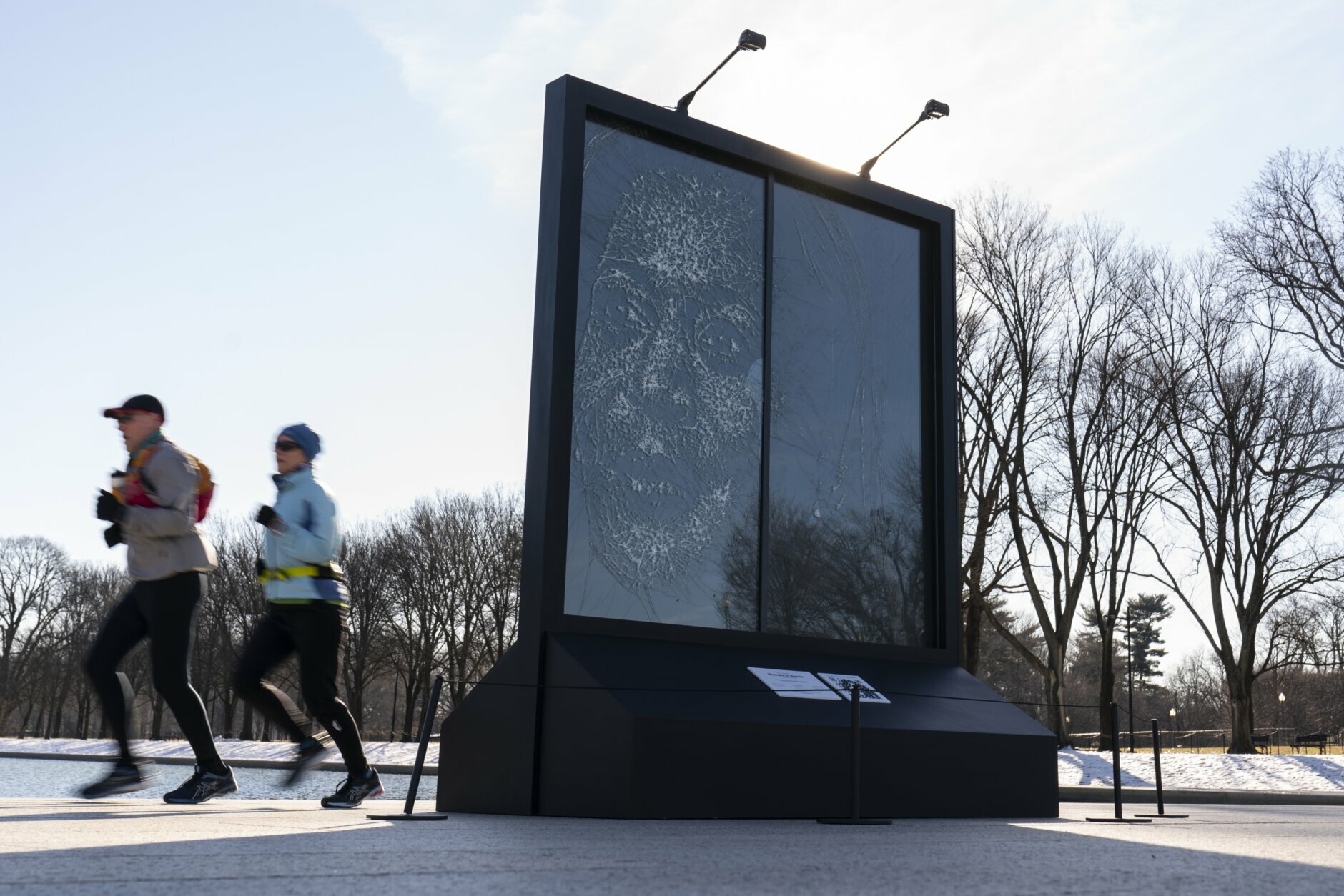 Joggers pass behind the installation "Vice President Kamala Harris Glass Ceiling Breaker" at the Lincoln Memorial in Washington, Wednesday, Feb. 4, 2021. Vice President Kamala Harris' barrier-breaking career has been memorialized in a portrait depicting her face emerging from the cracks in a massive sheet of glass. Using a photo of Harris that taken by photographer Celeste Sloman, artist Simon Berger lightly hammered on the slab of laminated glass to create the portrait of Harris. (AP Photo/Carolyn Kaster)