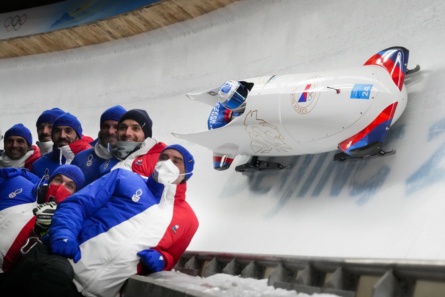 <p>Members of the Czech Republic team pose for a photo as teammates Dominik Dvorak and Jakub Nosek slide past during the 2-man heat 1 at the 2022 Winter Olympics, Monday, Feb. 14, 2022, in the Yanqing district of Beijing.</p>
