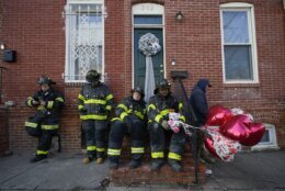 Baltimore City Fire emergency vehicle driver Jamaal Booker, second from right, holds balloons given to him by neighbor Darlene Cucina as a group of fire officials, from left, James Lamartina, Brian Petz, Justin Higley and Chris Hudson stand on a stoop across the street where three firefighters died in a building collapse while battling a two-alarm blaze in a vacant row home , Monday, Jan. 24, 2022, in Baltimore. Leaders in St. Louis and Baltimore are considering changes in dealing with fires at vacant homes after firefighters in both cities died at abandoned structures in January.  (AP Photo/Julio Cortez, File)