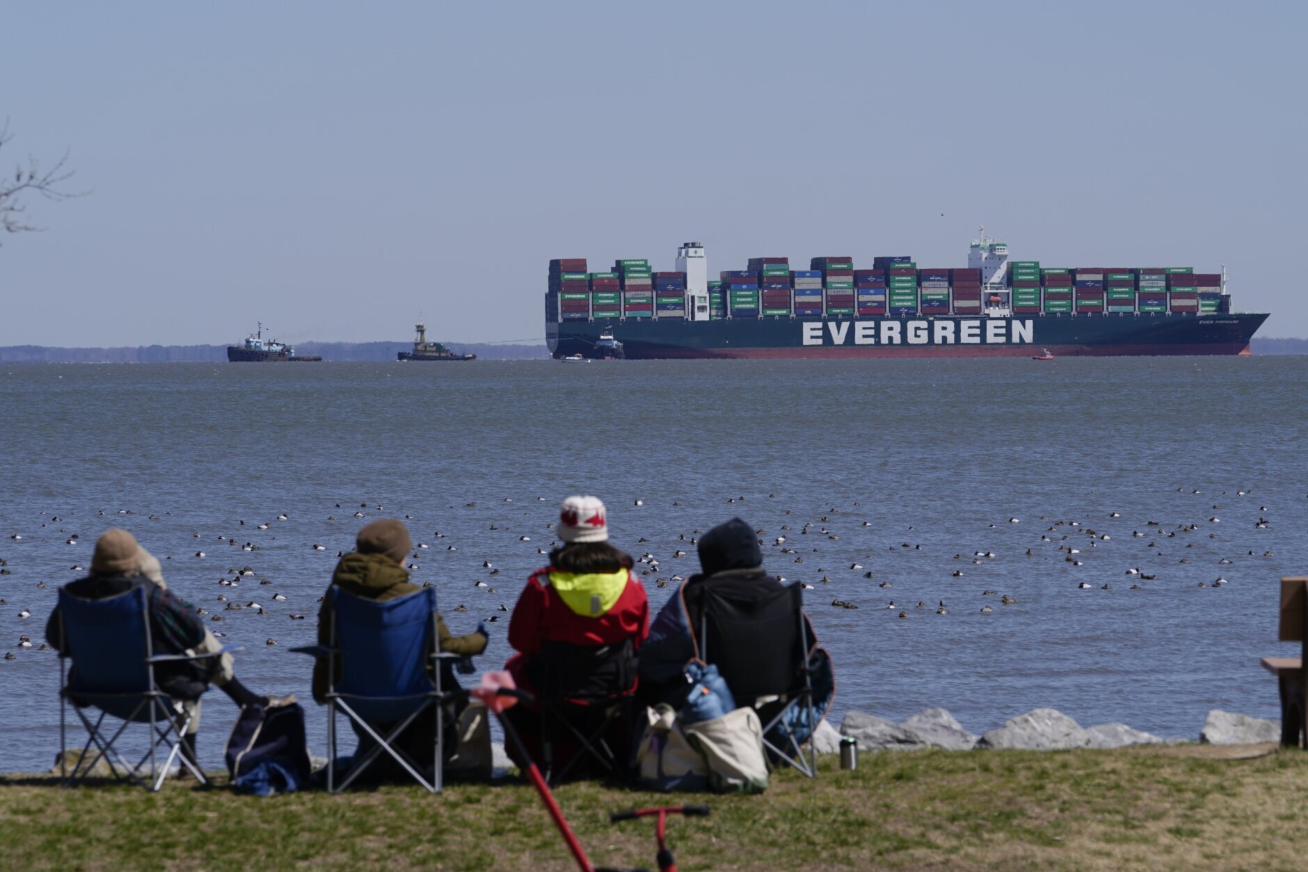 People sit in a park as they watch tugboats, top left, use lines to tow the container ship Ever Forward, top right, which ran aground in the Chesapeake Bay when crews Tuesday, March 29, 2022, attempted to refloat the ship in Pasadena, Maryland (AP Photo/Julio Cortez)