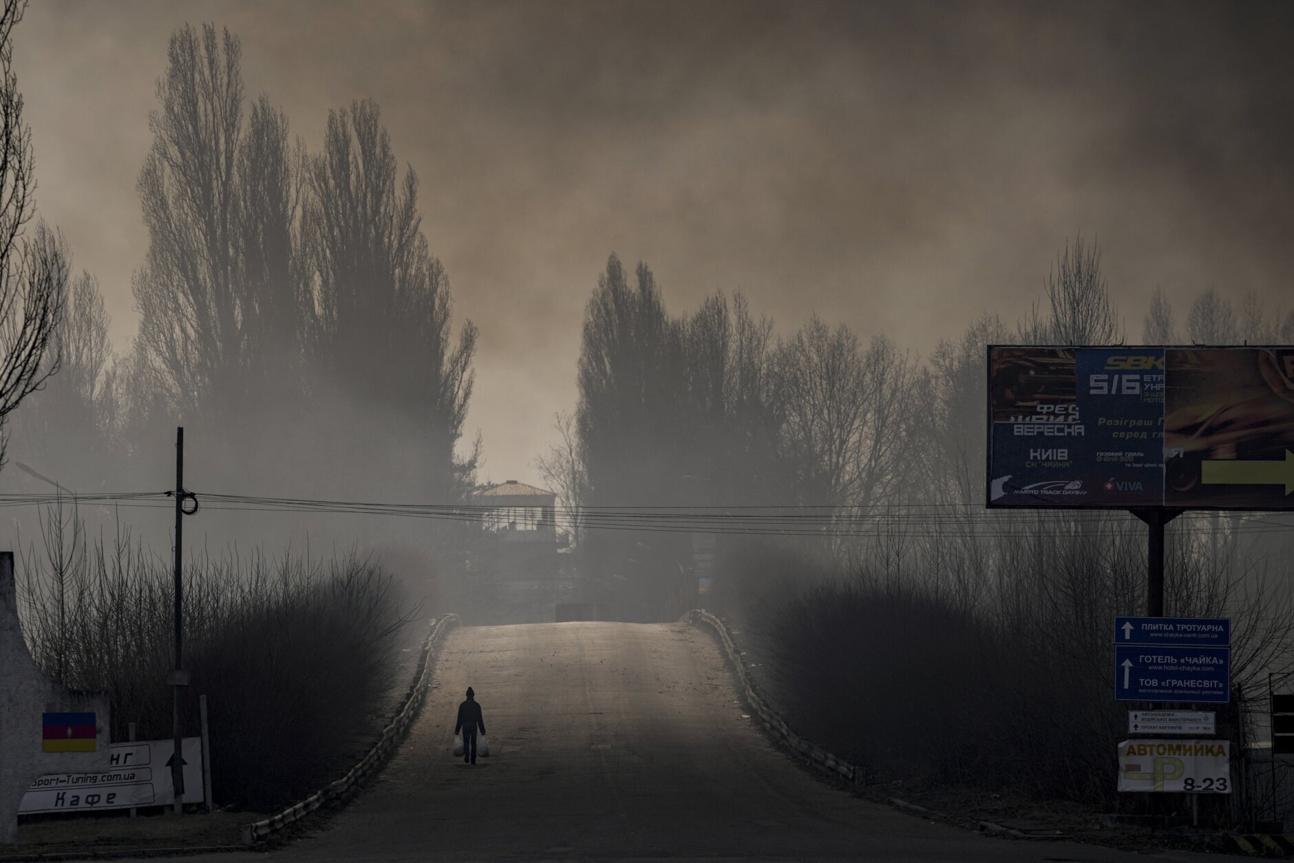 A man carries shopping bags as heavy smoke from a warehouse destroyed by Russian bombardment casts shadows on the road outside Kyiv, Ukraine,Thursday, March 24, 2022. (AP Photo/Vadim Ghirda)
