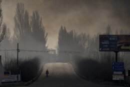 A man carries shopping bags as heavy smoke from a warehouse destroyed by Russian bombardment casts shadows on the road outside Kyiv, Ukraine,Thursday, March 24, 2022. (AP Photo/Vadim Ghirda)
