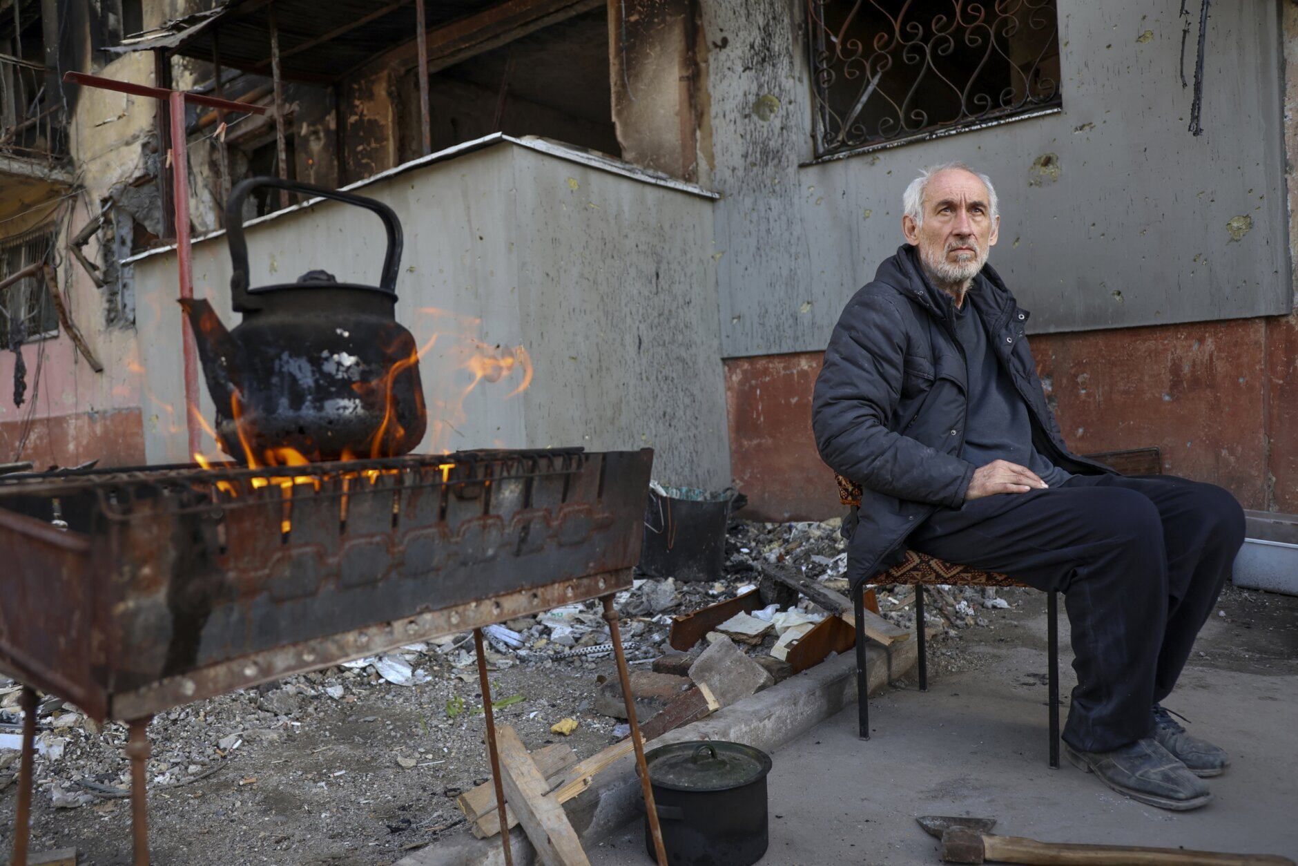 A local man sits in front of an apartment building damaged from heavy fighting as he waits for the kettle to boil in an area controlled by Russian-backed separatist forces in Mariupol, Ukraine, Tuesday, April 26, 2022. (AP Photo/Alexei Alexandrov)