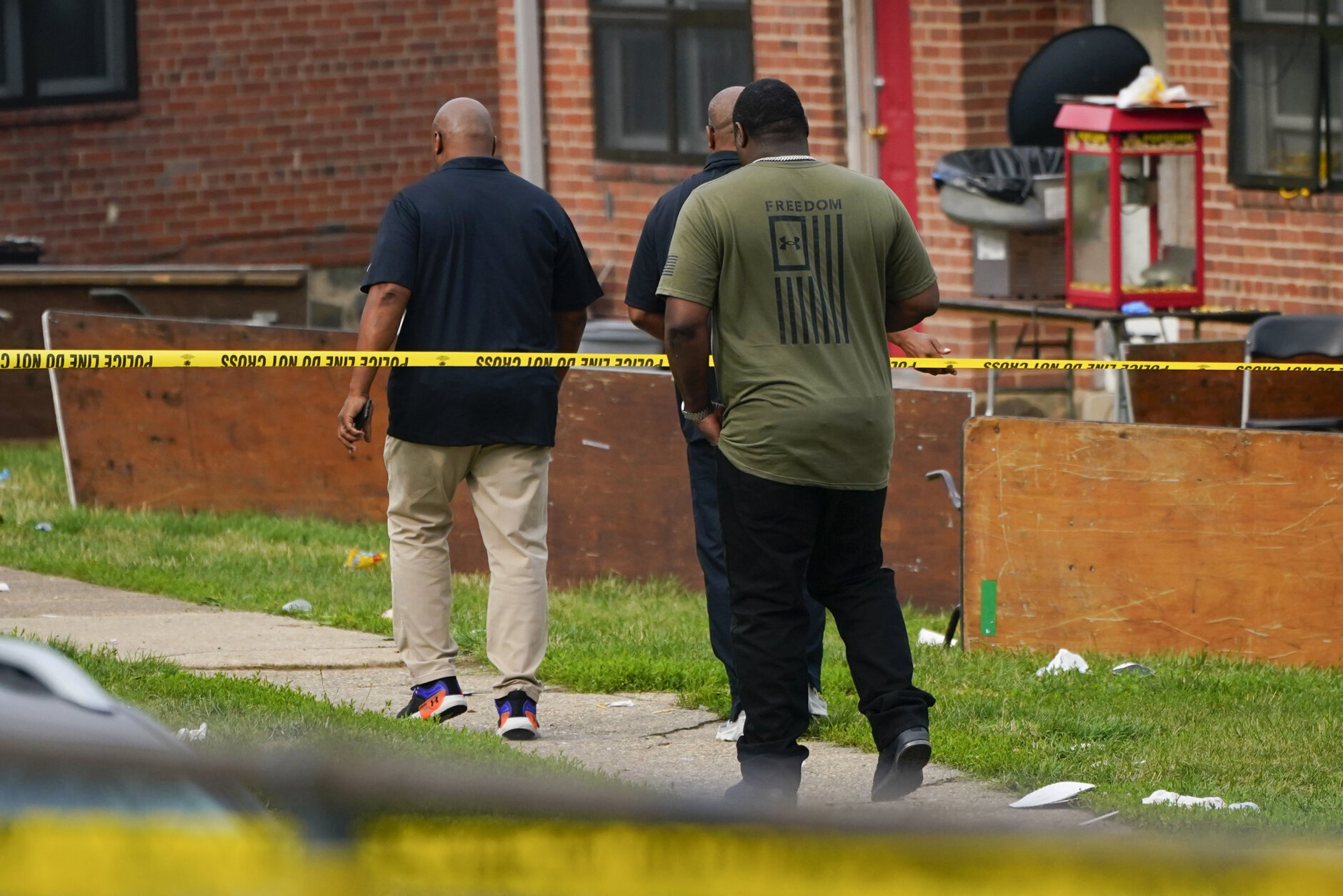 Members of the Baltimore Housing Authority walk near tables left on their side in the area of a mass shooting incident in the Southern District of Baltimore, Sunday, July 2, 2023. Police say a number of people were killed and dozens were wounded in a mass shooting that took place during a block party just after midnight. (AP Photo/Julio Cortez)