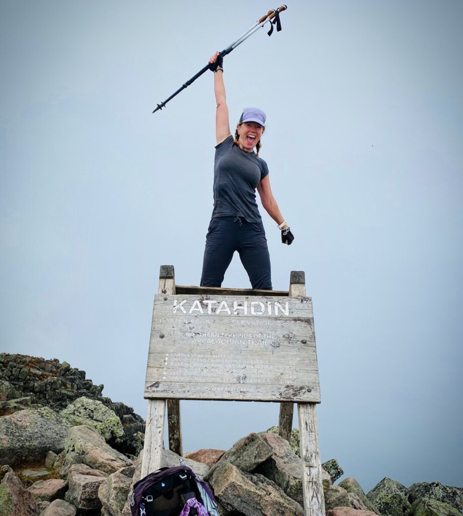 Cris Howard smiles widely with a fist pumped in the air in front of a sign at the top of Baxter's Peak on Mount Katahdin in Maine