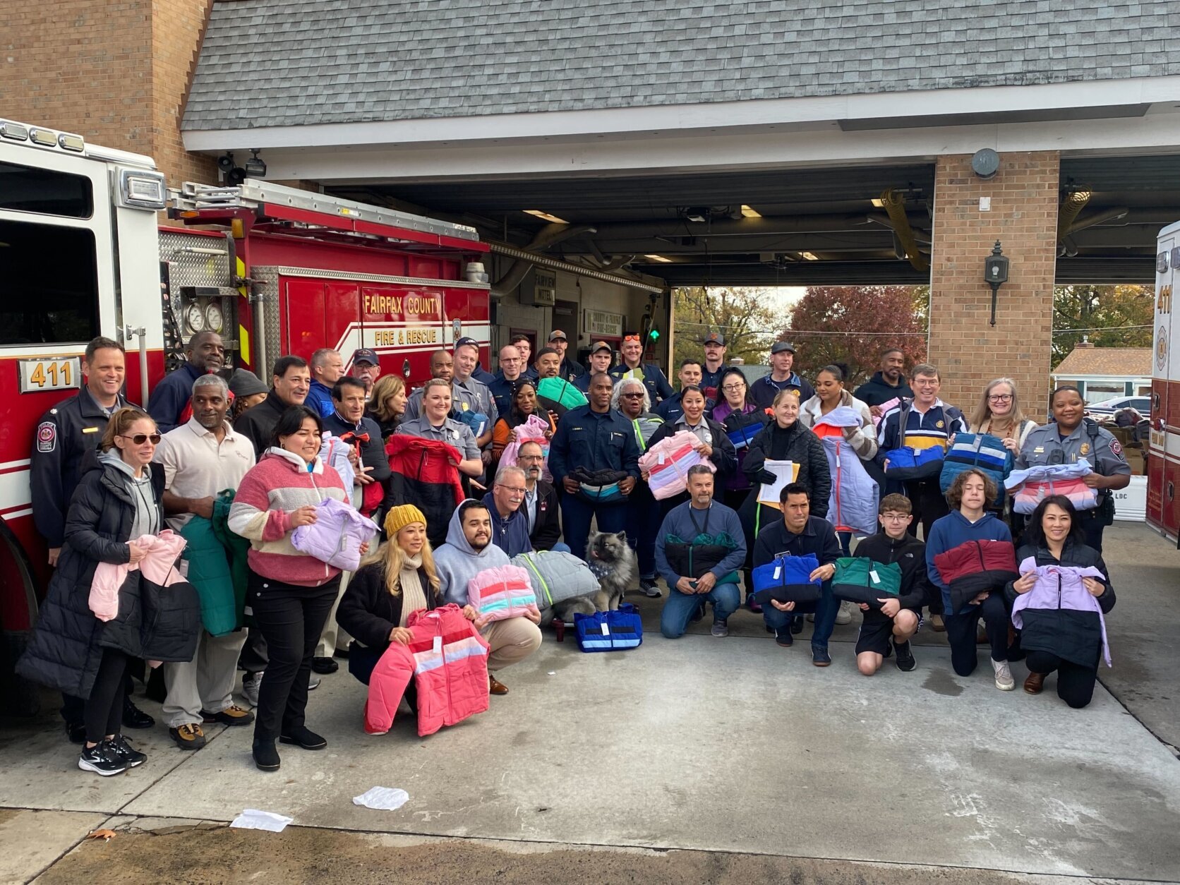 A crowd of people stand outside the firehouse holding up the free coats they received