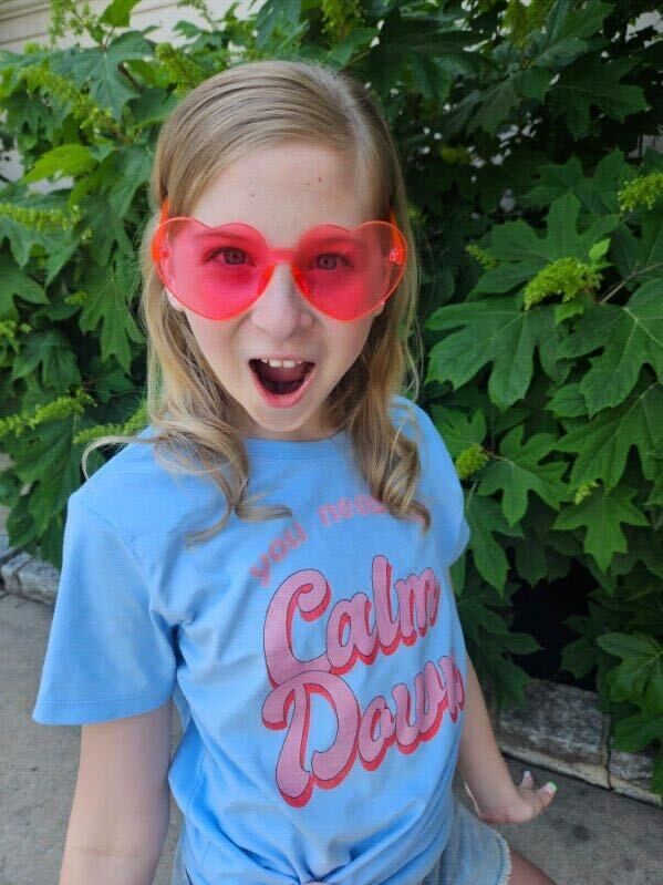 A young girl wears heart shaped glasses and a shirt that says "You Need to Calm Down."