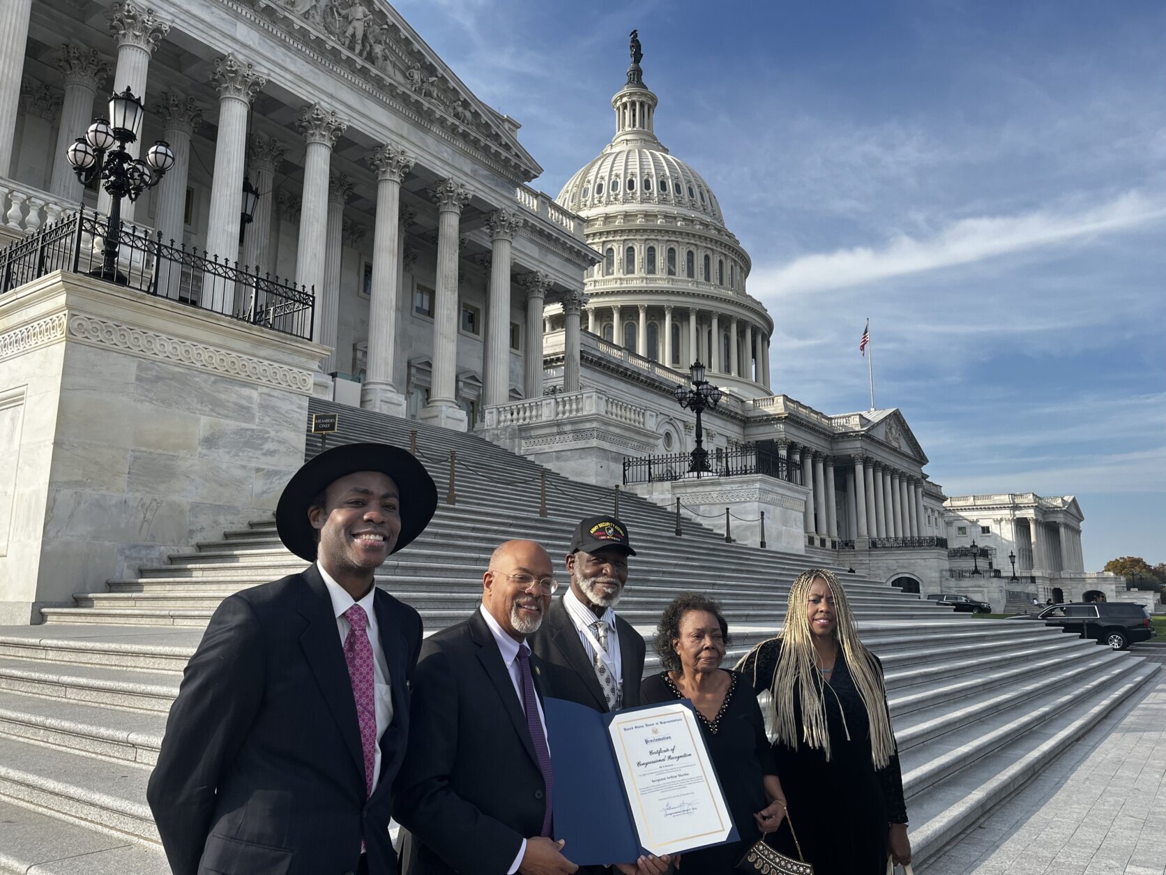 Sergeant Arthur Lee Martin stands at the steps of the U.S. Capitol Building in Washington, D.C. wearing a hat, just behind by four congressional leaders and staff members of color. Congressman Glenn Ivey stands in front of him holding a blue folder with a Congressional Citation inside.