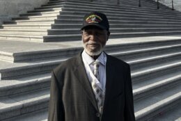 Sergeant Arthur Lee Martin stands at the bottom of the U.S. Capitol's front staircase wearing a black Army Security Agency baseball cap. Under a charcoal gray, lightly patterned suit rest a slightly obscured army badge just above a boxy-floral patterned crème colored tie and pin stripe button up shirt.