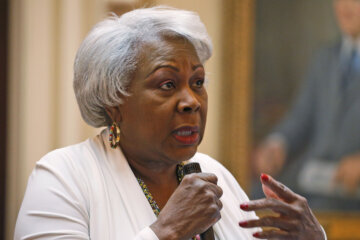 a woman in a white suit and gold earrings speaks into a microphone while gesticulating with her left arm and hand.