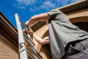 Young Male Property Inspector Climbing a Ladder and Photographing a gutter and roof shingles on a modern brown stucco home