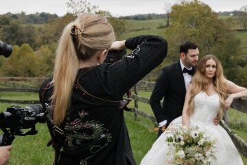On their wedding day, a couple leans against a fence in a grassy area as Jeanette takes their photo.