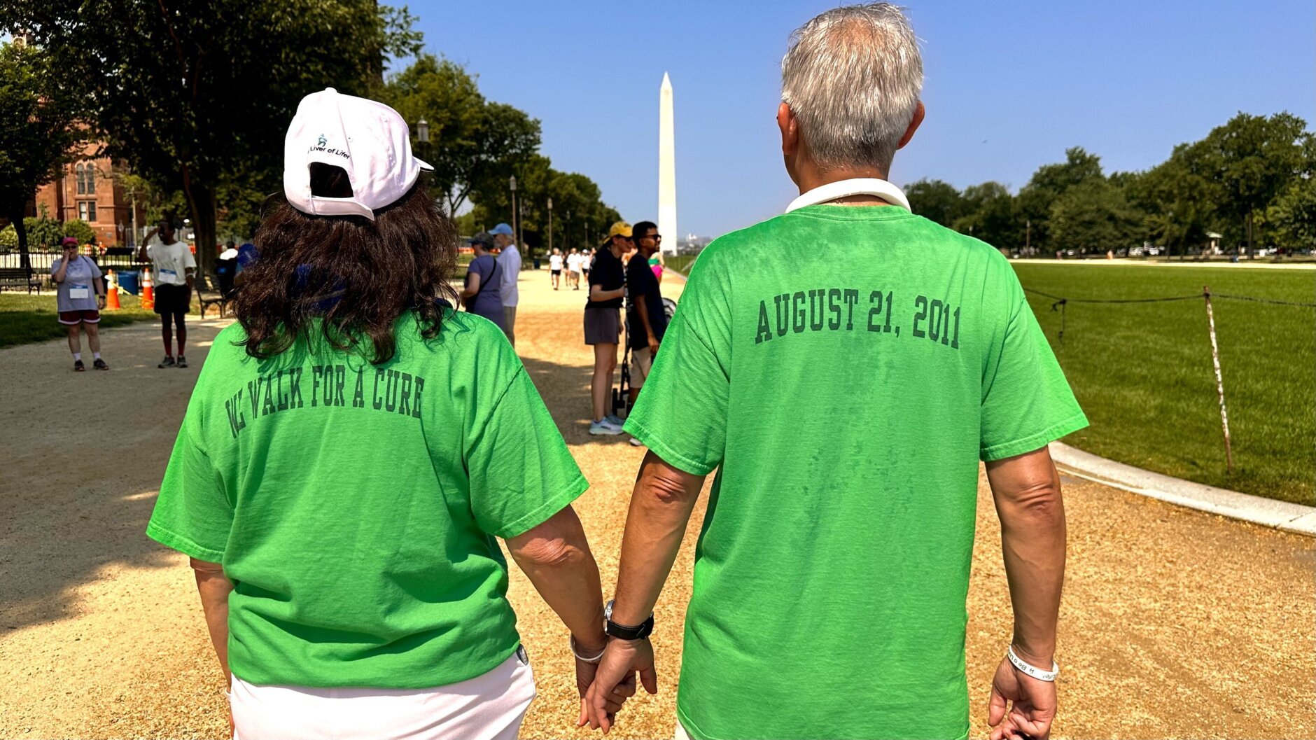 Da esquerda para a direita, uma mulher e um homem vestindo camisas verdes ficam de frente para um distante Monumento a Washington em um dia claro