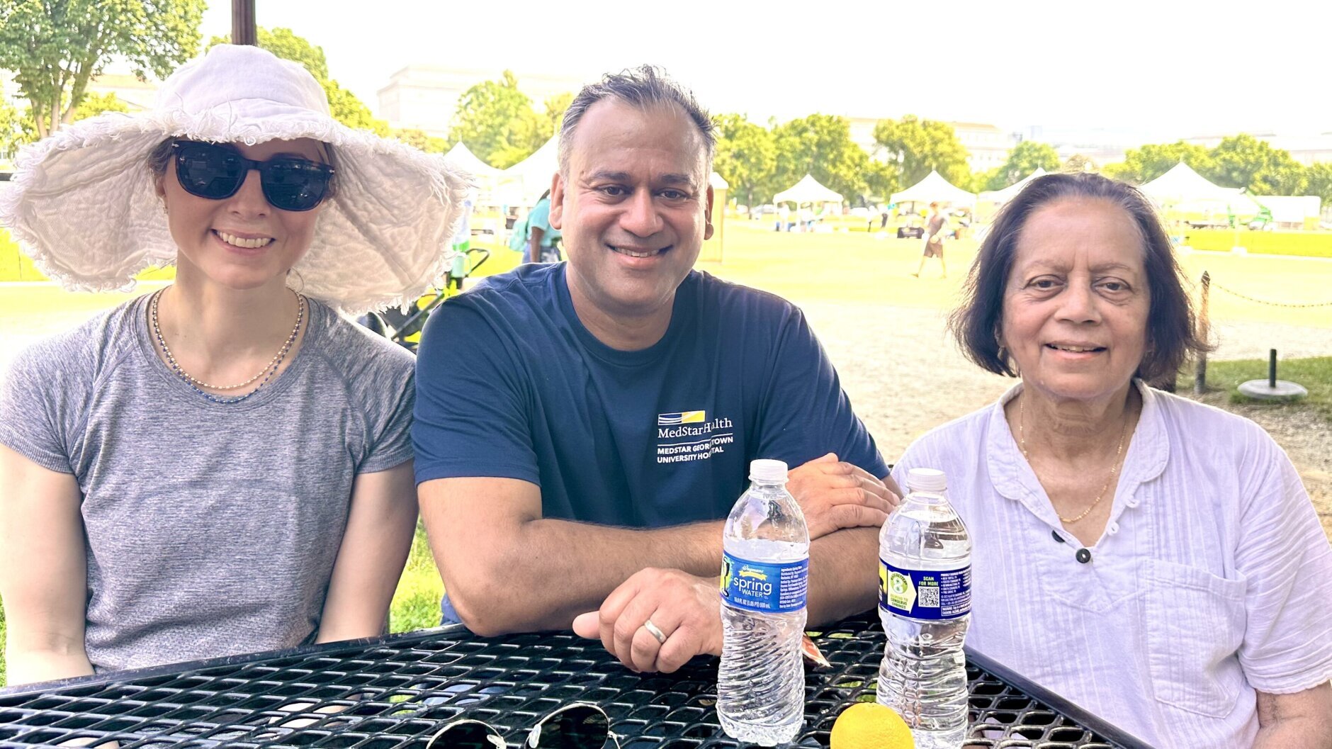 Dr. Rohit Satoskar (center) is seen at a table with his wife and mother to either side. One of the women is wearing a large sunhat while the other is positioned behind two water bottles.