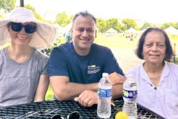 Dr. Rohit Satoskar (center) is seen at a table with his wife and mother to either side. One of the women is wearing a large sunhat while the other is positioned behind two water bottles.