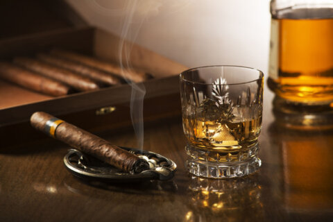 Glass of whiskey with ice cubes and smoking cigar on wooden table
