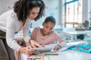 Female teacher helping young girl, teaching creative work in book and children in sunny classroom. Education empowerment in school, educator speaking to student and learning mathematics at desk