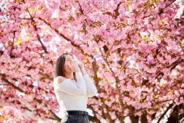 woman blows her nose near some cherry blossom trees