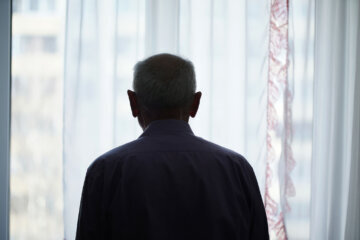 Silhouette of retired man looking through window with transparent curtain standing at home rear view. Loneliness and old human care concept.
