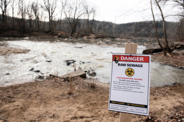 A recently placed warning sign is seen at the sight of a massive pipe rupture, as sewage flows into the Potomac River, right, in Glen Echo, Md., Friday, Jan. 23, 2026. A massive pipe that moves millions of gallons of sewage has ruptured and sent wastewater flowing into the Potomac River northwest of Washington, polluting it ahead of a major winter storm that has repair crews scrambling. (AP Photo/Cliff Owen)