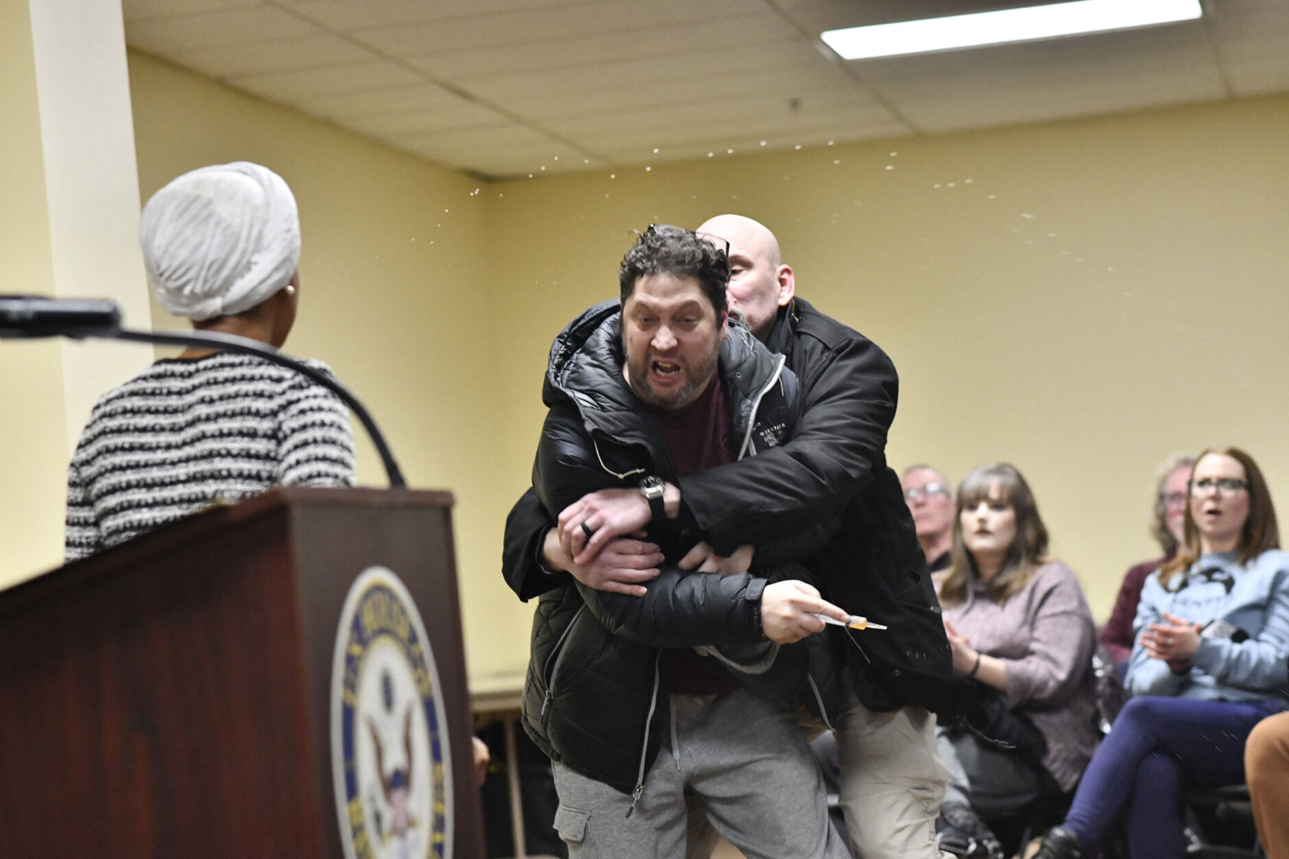 A man is tackled after spraying an unknown substance at US Representative Ilhan Omar (D-MN) (L) during a town hall she was hosting in Minneapolis, Minnesota, on January 27, 2026. US Democratic congresswoman Ilhan Omar was targeted during a speech late on January 27 by a man who sprayed an unidentified liquid at her from a syringe before being tackled by security guards, according to an AFP journalist at the scene. The man was led out of the premises as Omar, a frequent target of attacks by President Donald Trump, continued her speech saying "we will stay resilient in the face of whatever they might throw on us." (Photo by Octavio JONES / AFP via Getty Images)