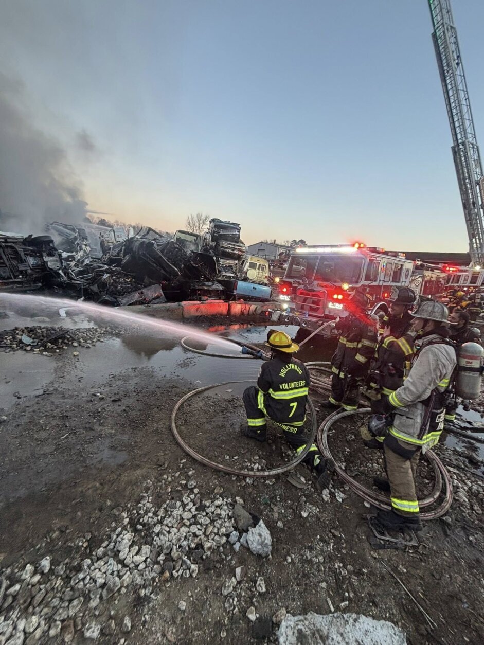 firefighters in junkyard aiming hose at fire to the left