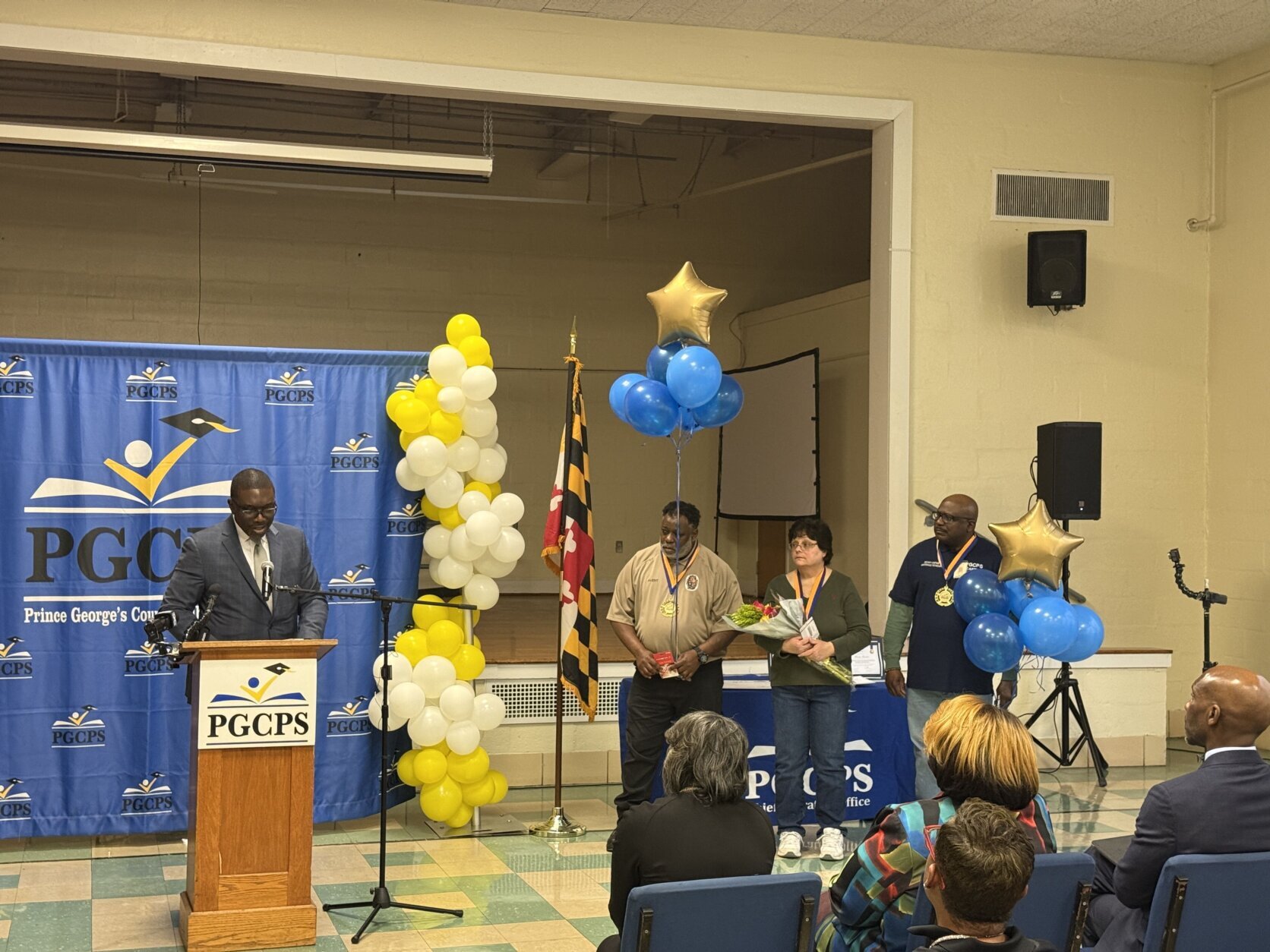 A man standing behind a podium, next to a column of balloons, the Maryland flag, and three honorees