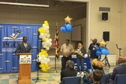 A man standing behind a podium, next to a column of balloons, the Maryland flag, and three honorees