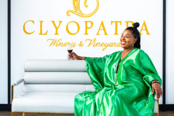 woman sitting on couch in green dress holding class of wine with winery sign in background