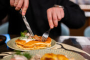 A person cuts into a stack of savory pancakes topped with sour cream and bacon with a fork and knife in a restaurant setting