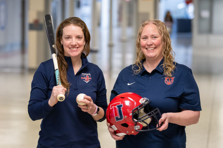 two women stand side by side one holding football helmet other holding baseball and bat