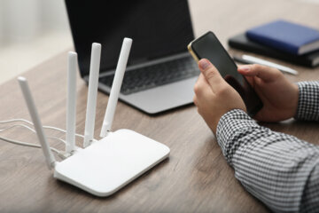 man hands holding smartphone next to internet router with laptop in the background