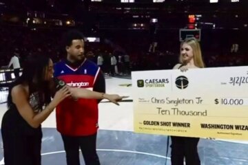 A woman points toward a large check held by another woman on the court. Christopher Singleton stands between them in a Wizards jersey