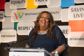 woman speaks at podium with sponsorship logos in background