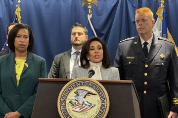 woman speaks at podium during news conference with other officials standing on either side