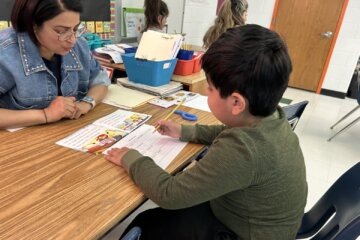Student works at table with his teacher Nuril Kingsley across