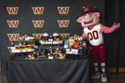 Washington Commanders' mascot, a giant anthropomorphic pig, poses to viewers' right of concessions arranged on a table at a press conference